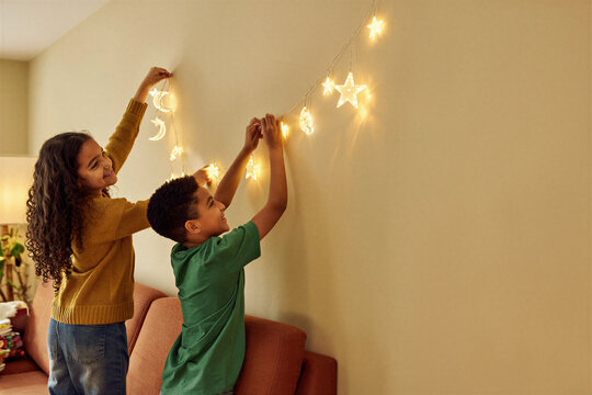 Two smiling siblings hanging crescent and star lights at home