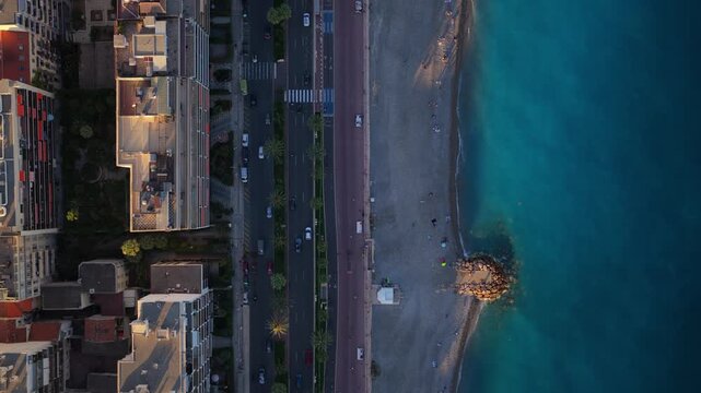 Top down flight along the Promenade des Anglais, palm lined road, pebble beach, groyne and turquoise sea in evening light, Nice, French Riviera, Cote d Azur, France