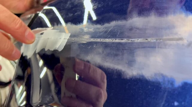 Professional auto mechanic man hands applying polyester car body filler putty with a steel spatula knife tool on a damaged blue vehicle part inside a detailing studio. Professional vehicle bodywork. 