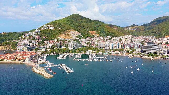 Budva, Montenegro rises above Adriatic marina, with dense waterfront buildings framed by green mountain slopes. Aerial panorama tracks harbor, boats and sunlit coastal scenery beneath clouds.