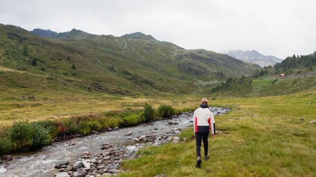 Static wide shot of a hiker walking away along a rocky mountain stream in a green alpine valley on an overcast day, Dolomites, South Tyrol, Alps, Italy