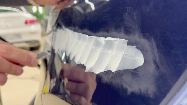 Professional auto mechanic man hands applying polyester car body filler putty with a steel spatula knife tool on a damaged blue vehicle part inside a detailing studio. Professional vehicle bodywork. 