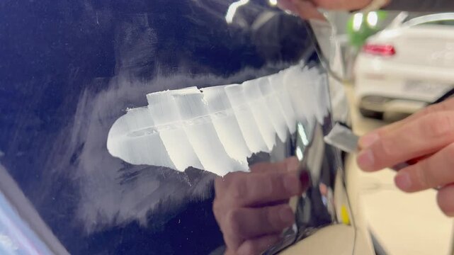 Professional auto mechanic man hands applying polyester car body filler putty with a steel spatula knife tool on a damaged blue vehicle part inside a detailing studio. Professional vehicle bodywork. 