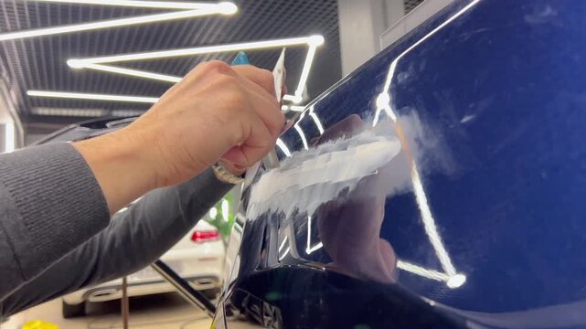 Professional auto mechanic man hands applying polyester car body filler putty with a steel spatula knife tool on a damaged blue vehicle part inside a detailing studio. Professional vehicle bodywork. 