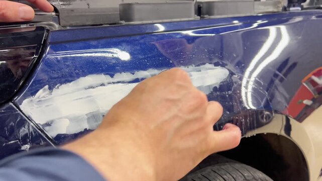 Professional car detailer hands using a sanding block and sandpaper tool for wet sanding body filler on a blue metallic automobile fender. Vehicle bodywork repair, paint preparation, and restoration. 