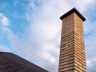 Fototapeta premium Tall chimney built from fireclay bricks rising above a shingle roof against blue cloudy sky. Traditional masonry chimney for stove, fireplace and home heating.