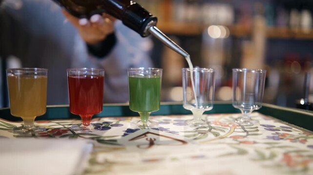 The bartender pours different liqueurs into small shot glasses. The liqueurs are of different colors. Close‑up shot.