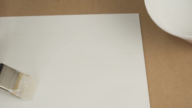 Gloved hand using a paintbrush to apply white paint or primer to a sheet of paper or board