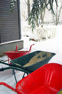 Bright wheelbarrows in snowy yard, one loaded with de-icing sand mixture, the other empty. Embodies ice hazard reduction, sustainable salt alternative, and urban park safety management during winter.