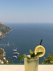Fototapeta premium Refreshing Lemonade Cocktail with Mint and Lemon Slice Overlooking Scenic Coastal Town and Ocean View