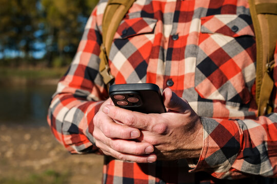 Man in plaid shirt using smartphone by lake during early autumn, holding mobile device outdoors against peaceful natural landscape, digital communication concept