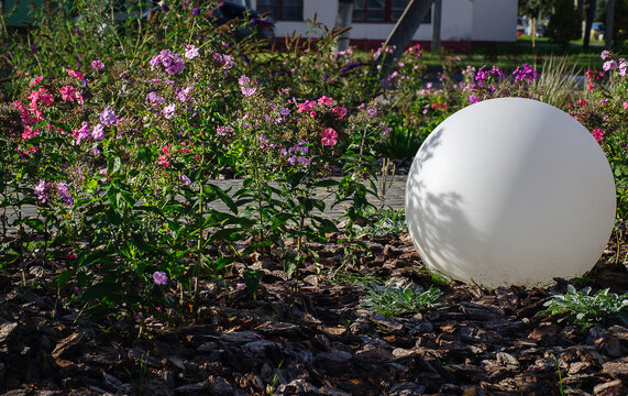 Large white round lamp standing on bark mulch next to blooming pink phlox flowers in landscaped public city park garden with copy space