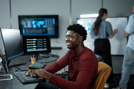 Young African male software engineer writing program code on multiple desktop screens while his colleagues working in the background in a modern office.