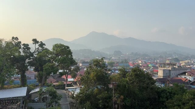 Hazy mountains loom behind clustered homes. Tropical trees frame the view. Red roofs dot the village below. Soft light blankets the entire scene. Quiet, peaceful morning in a mountain town