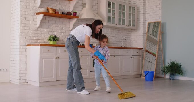 Caring young mother teaches her little daughter to clean using broom to sweep house. Mother in blue gloves demonstrates sweeping techniques, showing daughter how to effectively handle household chores
