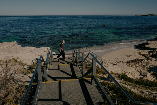 Bald man descending steps toward a sandy beach with clear blue w