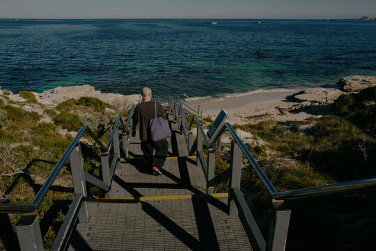Bald man descending steps toward a sandy beach with clear blue w