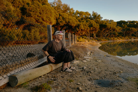 Smiling man in a beige cap sitting on a log by a lake with a quo