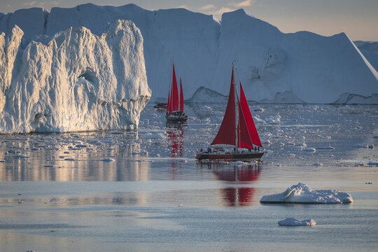 Red sailboats sailing past giant icebergs in arctic waters