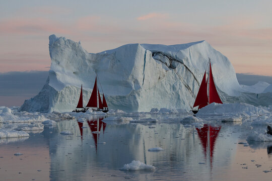 Sailboats with striking red sails near a massive iceberg at dusk