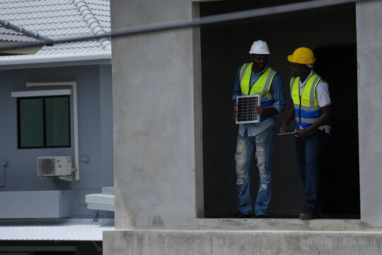 Two African engineers in safety vests and hard hats prepare the panel installation at the entrance, laptop in hand outdoors at a modern housing estate project with copy space