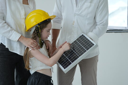 Happy young family with little daughter in a toy hard hat let the girl in a toy helmet touch the photovoltaic cells up close in their future eco friendly house, authentic everyday scene
