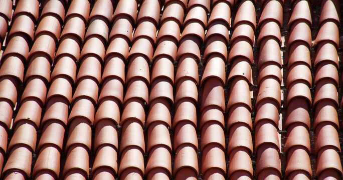 Close-up of traditional terracotta roof tiles arranged in a repeating pattern, showing warm red clay colors, curved shapes, and strong sunlight and shadows. Rustic geometric. texture roofing