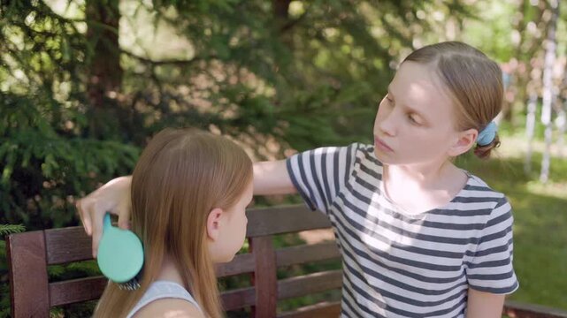 Two sisters share quiet grooming moment sitting together outdoors on park bench. Caring child carefully detangles long blonde hair of younger sibling using plastic hairbrush with gentle care.