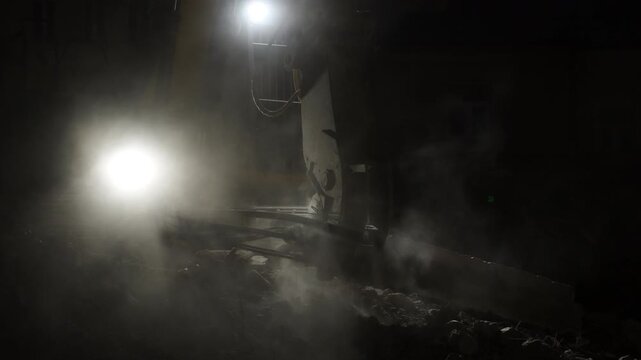 dramatic night scene inside an industrial or demolition site, a bright white work light glaring through thick dust and fog, a large mechanical arm or crane silhouette partially visible on the right