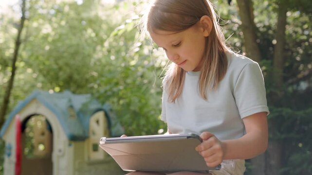 Engrossed child holds digital tablet while playing game in sunny backyard garden. Little girl experiences summer digital entertainment sitting outside surrounded by green summer nature.