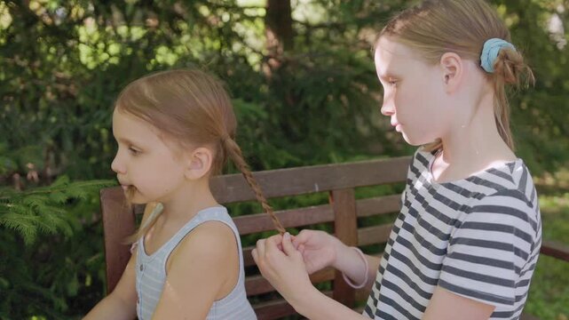 Young girl braids sister hair outdoors while seated on wooden garden bench, showing sibling conflict annoyance as younger child sits with bored impatient expression in sunny park setting.
