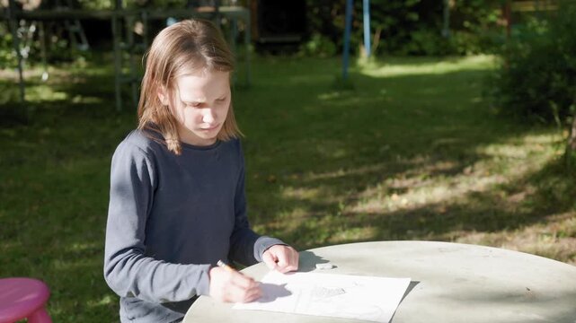 Young girl draws with pencil on paper at round stone table in sunny garden. Focused female child sketches outdoors during daytime, displaying thoughtful emotion while sitting on stool.