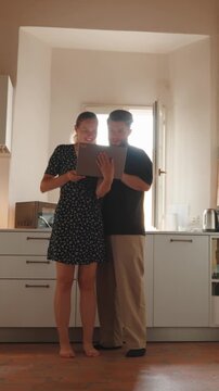 Happy young couple using a laptop computer for online shopping and planning their future together, smiling and laughing while standing in their modern bright kitchen. Vertical video