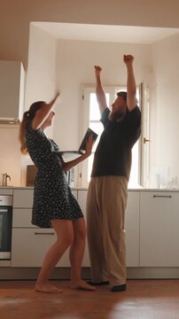Excited young couple celebrating good news on a laptop, dancing and hugging in the kitchen while sharing a moment of pure joy, happiness and success together at home. Vertical video