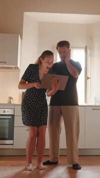 Overjoyed couple celebrating a successful online purchase or winning a prize on a laptop, dancing and high-fiving in their bright and modern kitchen at home. Vertical video