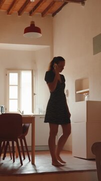 Young woman wearing a casual dress happily talking on the phone while walking barefoot around her cozy apartment, enjoying a sunny morning with a relaxed and cheerful attitude. Vertical video