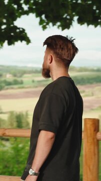 Young bearded man admiring a breathtaking green countryside panorama from a scenic viewpoint during a peaceful and relaxing travel journey on a beautiful sunny summer day. Vertical video 