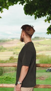 Young bearded man standing at wooden fence and contemplating the serene tuscan countryside, finding peace and inspiration in the picturesque landscape on a calm day. Vertical video 