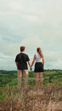 Young romantic couple holding hands and watching a beautiful green landscape while standing on a hill, enjoying the tranquility of nature and their loving relationship. Vertical video 