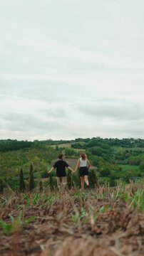Romantic young couple holding hands and running down a hill in the italian countryside, enjoying a slow living vacation and expressing freedom in their relationship. Vertical video 