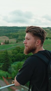 Contemplative bearded male backpacker admiring the serene beauty of the rolling green hills and landscape from a viewpoint, feeling a sense of freedom and wonder. Vertical video 