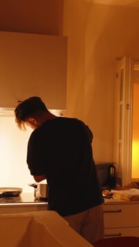 Silhouette of a young man cooking in the kitchen, stirring food in a pot on the stove during a warm sunset, creating a cozy and domestic home atmosphere. Vertical video 