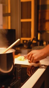 Close up of a man cooking a delicious meal in a cozy kitchen, chopping vegetables on a cutting board while water boils in a steaming pot on an induction hob. Vertical video 
