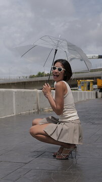 Asian woman crouches confidently holding a transparent umbrella on a city street, wearing a beige crop top, pleated mini skirt, and strappy heels
