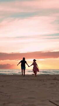 Romantic young couple running on a sandy beach holding hands at sunset, a serene summer vacation concept showing love, joy, and freedom in a beautiful tropical seaside setting. Vertical video