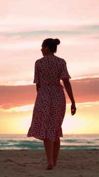 Woman in dress dancing barefoot on sandy beach, walking towards ocean at sunset, enjoying freedom, silhouette against colorful sky, carefree vacation moment. Vertical video