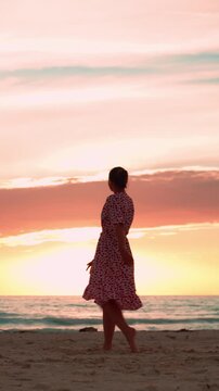 Silhouette of a happy woman dancing barefoot on the sandy beach against a beautiful and romantic sunset, enjoying her freedom and celebrating the summer vacation. Vertical video