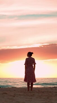 Woman wearing a dress standing on a sandy beach contemplating a stunning orange and pink sunset, enjoying a moment of tranquility, serenity, and mindfulness on her holiday. Vertical video