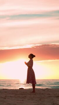 Silhouette of a spiritual woman practicing yoga and meditation while praying on a beautiful sandy beach with a peaceful and inspiring orange and pink sunset over the sea. Christian woman. Vertical