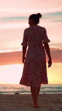 Carefree woman in a summer dress dancing barefoot on a sandy beach, enjoying a beautiful and romantic sunset over the calm ocean, representing freedom and tranquility. Vertical video